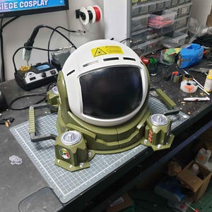 May include: A white and green helmet with a black visor and a warning sign on the top. The helmet is attached to a green base with four legs and a handle on each side.