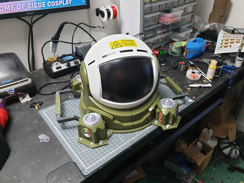 May include: A white and green helmet with a black visor and a warning sign on the top. The helmet is attached to a green base with four legs and a handle on each side.