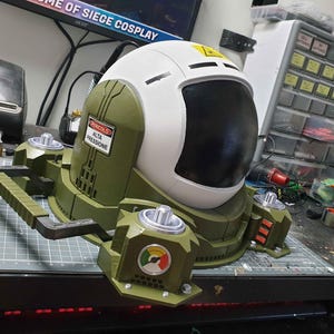 May include: A green and white helmet with a large black visor and a warning label that reads "PERICOLO ALTA PRESSIONE". The helmet is attached to a green and black base with a handle.