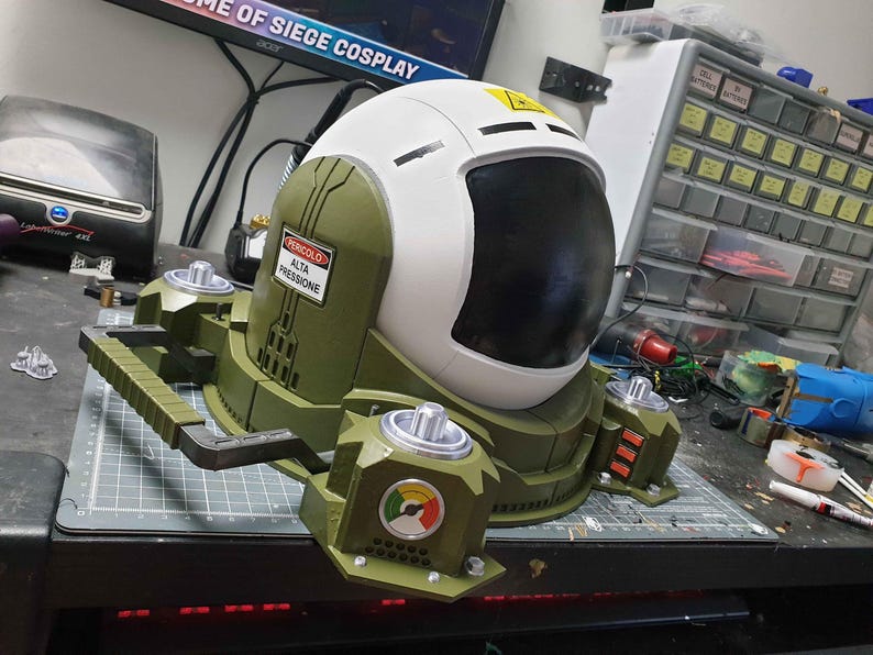 May include: A green and white helmet with a large black visor and a warning label that reads "PERICOLO ALTA PRESSIONE". The helmet is attached to a green and black base with a handle.