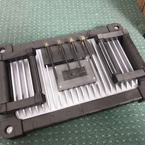May include: A silver and black metal box with two black handles on each side. The box has a series of small, black metal rods protruding from the top. The box has a black, rectangular label with a wavy line on it.