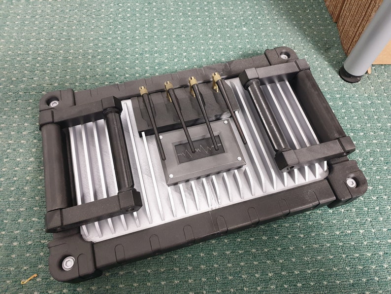 May include: A silver and black metal box with two black handles on each side. The box has a series of small, black metal rods protruding from the top. The box has a black, rectangular label with a wavy line on it.