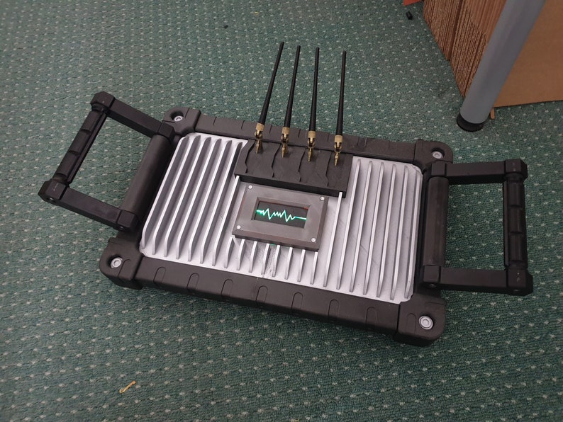 May include: A black and silver device with four antennas and a small screen displaying a green waveform. The device has two black handles on the sides.