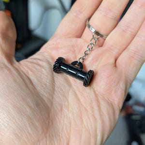 May include: A black keychain featuring a miniature cannon design. The cannon has a cylindrical body with detailed wheel-like ends and is attached to a silver chain and keyring. The keychain is held in a hand.