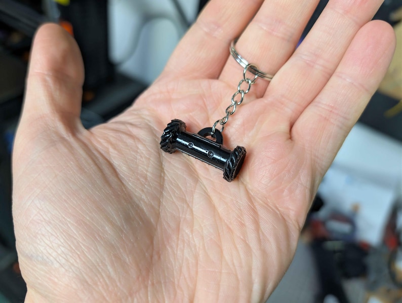 May include: A black keychain featuring a miniature cannon design. The cannon has a cylindrical body with detailed wheel-like ends and is attached to a silver chain and keyring. The keychain is held in a hand.