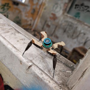 K&ouml;nnte beinhalten: Ein kleiner Roboter-Spinne in Beige und Braun, mit vier beweglichen Beinen und einem zentralen K&ouml;rper mit einem blau-gr&uuml;nen kreisf&ouml;rmigen Design. Der Hintergrund ist eine verwitterte, mit Graffiti bedeckte Oberfl&auml;che.