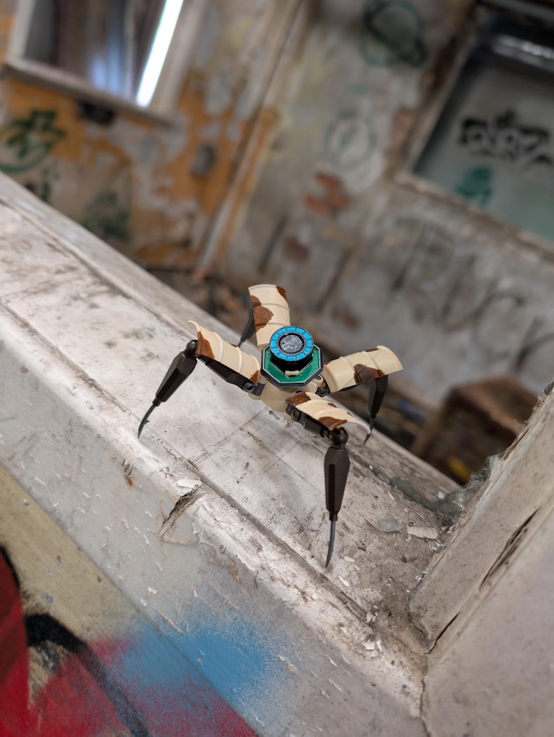 K&ouml;nnte beinhalten: Ein kleiner Roboter-Spinne in Beige und Braun, mit vier beweglichen Beinen und einem zentralen K&ouml;rper mit einem blau-gr&uuml;nen kreisf&ouml;rmigen Design. Der Hintergrund ist eine verwitterte, mit Graffiti bedeckte Oberfl&auml;che.