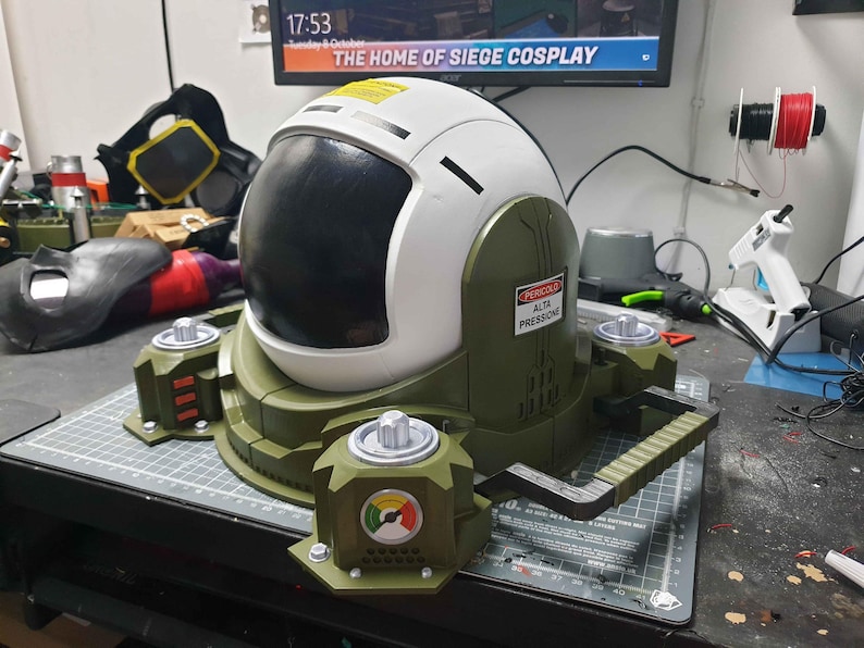 May include: A white and green helmet with a black visor and a warning label that reads "PERICOLO ALTA PRESSIONE". The helmet is attached to a green and black base with a handle.