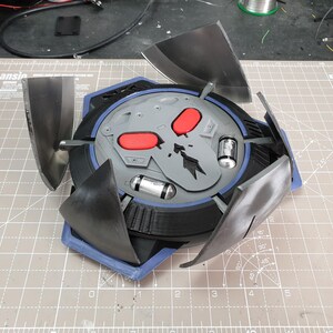 May include: A gray and black 3D printed model of a weapon from a video game. The weapon has a skull design with red eyes and four black blades that can be extended.