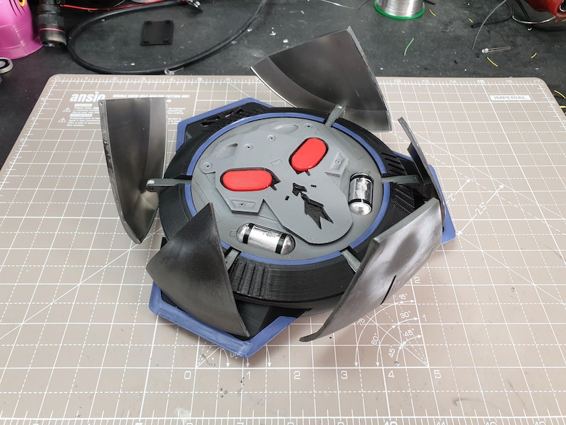 May include: A gray and black 3D printed model of a weapon from a video game. The weapon has a skull design with red eyes and four black blades that can be extended.