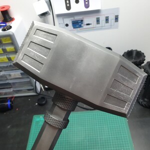 May include: A gray plastic hammer with a textured handle and a flat, rectangular head. The head has a series of raised lines on the top and bottom.