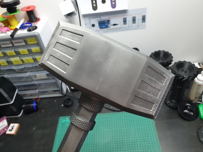 May include: A gray plastic hammer with a textured handle and a flat, rectangular head. The head has a series of raised lines on the top and bottom.