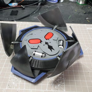 May include: A 3D printed, black and gray, skull-shaped object with red eyes. The object has four black, triangular wings that are folded down. The object is sitting on a gray cutting mat.