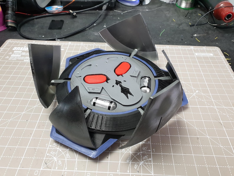 May include: A 3D printed, black and gray, skull-shaped object with red eyes. The object has four black, triangular wings that are folded down. The object is sitting on a gray cutting mat.