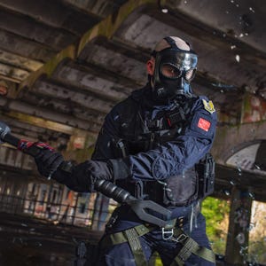 May include: A person wearing a black tactical vest and a gas mask holds a black and red sledgehammer in a dark, industrial setting.