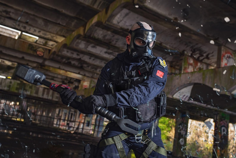 May include: A person wearing a black tactical vest and a gas mask holds a black and red sledgehammer in a dark, industrial setting.