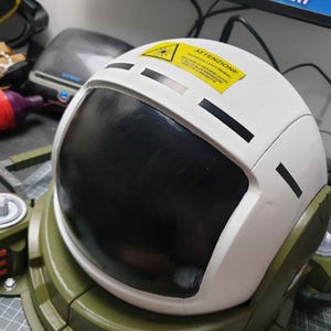 May include: A white astronaut helmet with a black visor and a yellow warning label that reads "ATTENZIONE!"  The helmet is attached to a green and black base.