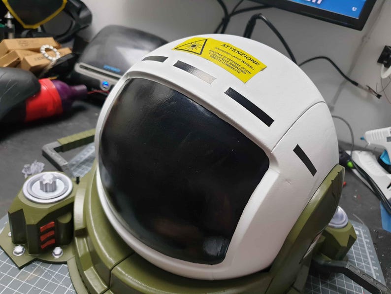 May include: A white astronaut helmet with a black visor and a yellow warning label that reads "ATTENZIONE!"  The helmet is attached to a green and black base.