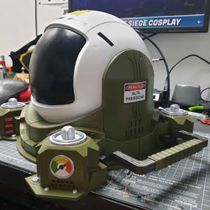 May include: A white and green helmet with a black visor and a warning sign that reads "Pericolo Alta Pressione". The helmet is attached to a green and black base with a handle.