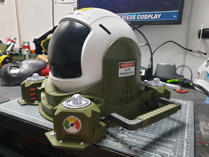 May include: A white and green helmet with a black visor and a warning sign that reads "Pericolo Alta Pressione". The helmet is attached to a green and black base with a handle.