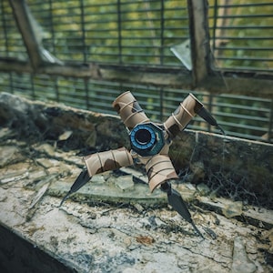 K&ouml;nnte beinhalten: Ein futuristisches, mechanisches Ger&auml;t mit vier Beinen und einem zentralen Hub. Die Beine sind dunkel mit scharfen Spitzen, und der K&ouml;rper weist ein Tarnmuster auf. Der zentrale Hub hat ein blaues und schwarzes kreisf&ouml;rmiges Design. Das Ger&auml;t befindet sich vor einem unscharfen Hintergrund.