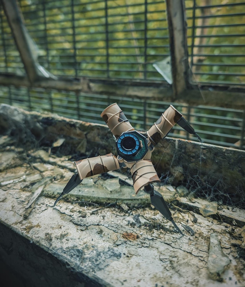 K&ouml;nnte beinhalten: Ein futuristisches, mechanisches Ger&auml;t mit vier Beinen und einem zentralen Hub. Die Beine sind dunkel mit scharfen Spitzen, und der K&ouml;rper weist ein Tarnmuster auf. Der zentrale Hub hat ein blaues und schwarzes kreisf&ouml;rmiges Design. Das Ger&auml;t befindet sich vor einem unscharfen Hintergrund.