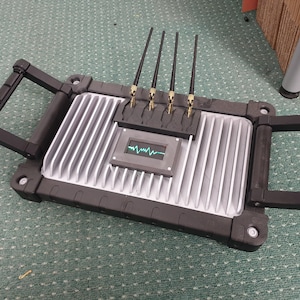 May include: A black and silver device with four antennas and a small screen displaying a green waveform. The device has two black handles on the sides.