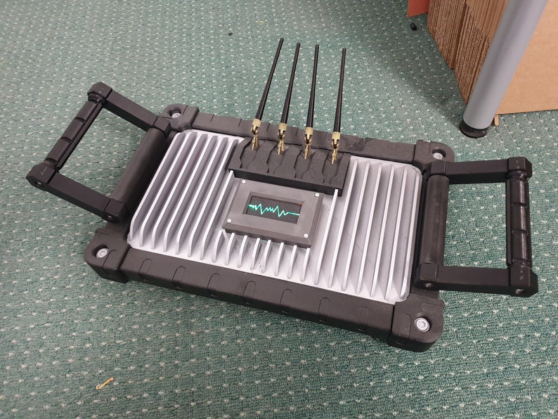 May include: A black and silver device with four antennas and a small screen displaying a green waveform. The device has two black handles on the sides.