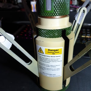 May include: A tan and green high voltage entry-denial device with a warning label. The label reads "Danger Electrical shock risk". The device has metallic arms extending outwards.