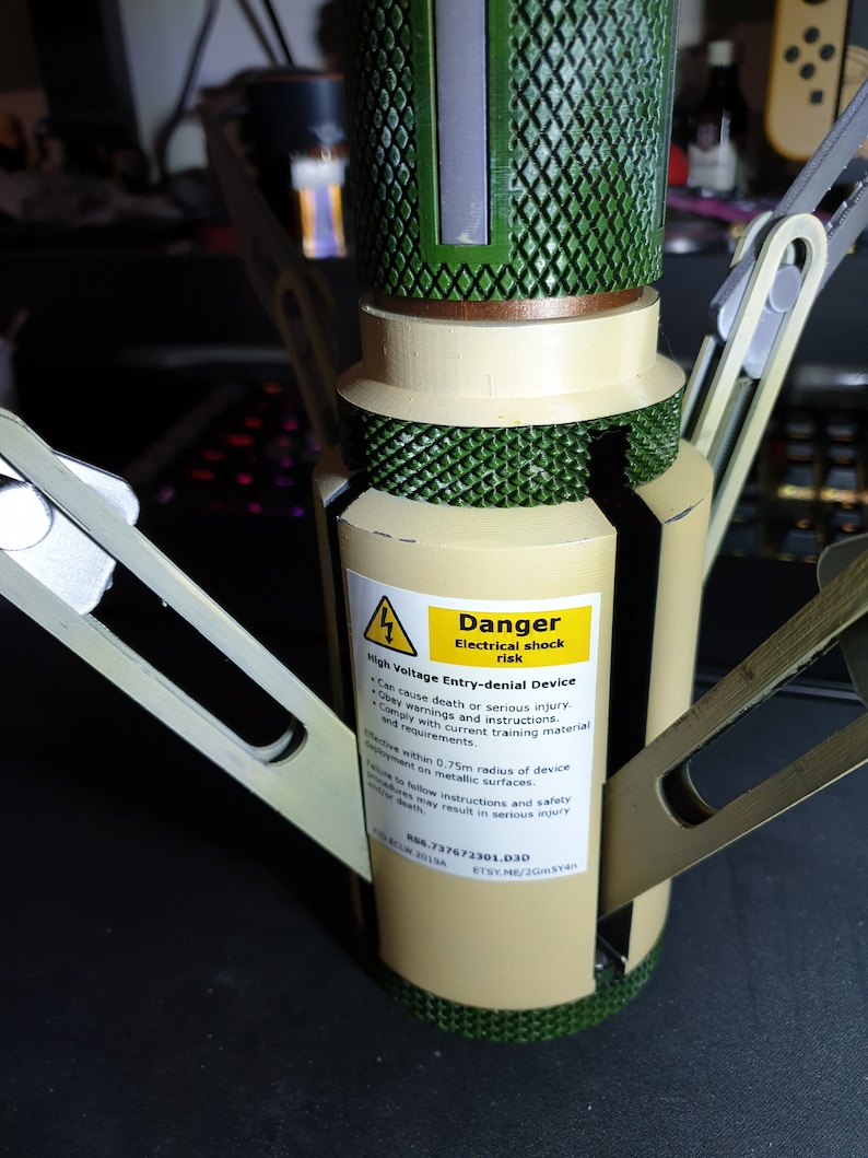 May include: A tan and green high voltage entry-denial device with a warning label. The label reads "Danger Electrical shock risk". The device has metallic arms extending outwards.