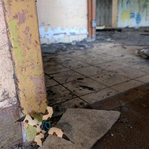 K&ouml;nnte beinhalten: Eine kleine Drohne mit mehreren Rotoren, einer zentralen Kamera und vier Armen, jeweils mit einem Propeller. Die Drohne ist beige, wei&szlig; und braun. Sie liegt auf einem Betonst&uuml;ck in einem bauf&auml;lligen Raum mit abbl&auml;tternder Farbe und Graffiti.