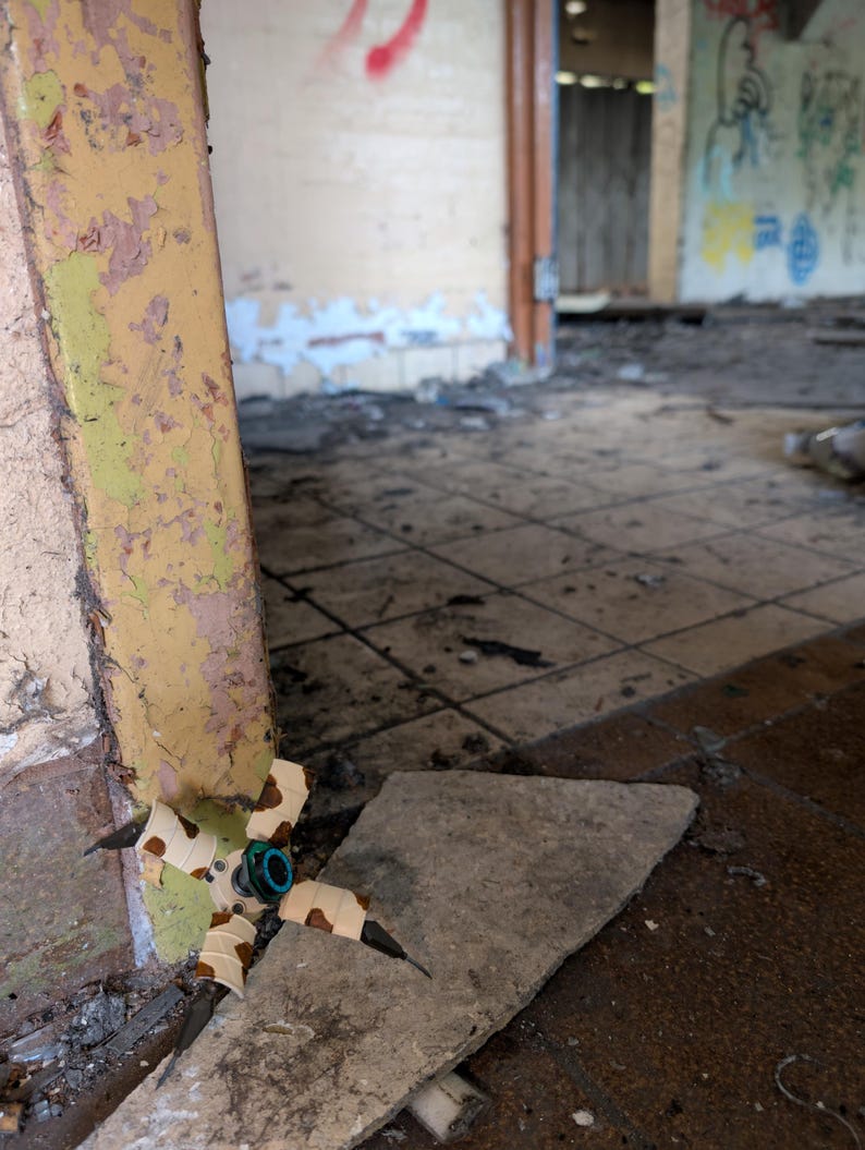 K&ouml;nnte beinhalten: Eine kleine Drohne mit mehreren Rotoren, einer zentralen Kamera und vier Armen, jeweils mit einem Propeller. Die Drohne ist beige, wei&szlig; und braun. Sie liegt auf einem Betonst&uuml;ck in einem bauf&auml;lligen Raum mit abbl&auml;tternder Farbe und Graffiti.