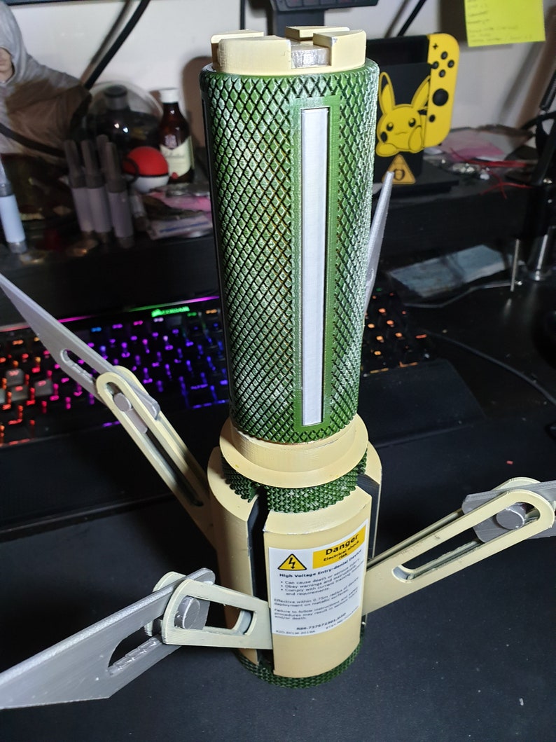 May include: A 3D-printed device with a green, diamond-patterned cylindrical body and beige accents. It features a white vertical stripe and extending gray arms. A yellow sticker with a lightning bolt and the word "Danger" is visible.