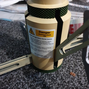 May include: A beige and green cylindrical device with a warning label that reads "Danger Electrical shock risk." The label also states "High Voltage Entry-denial Device." The device has a textured green top and base.