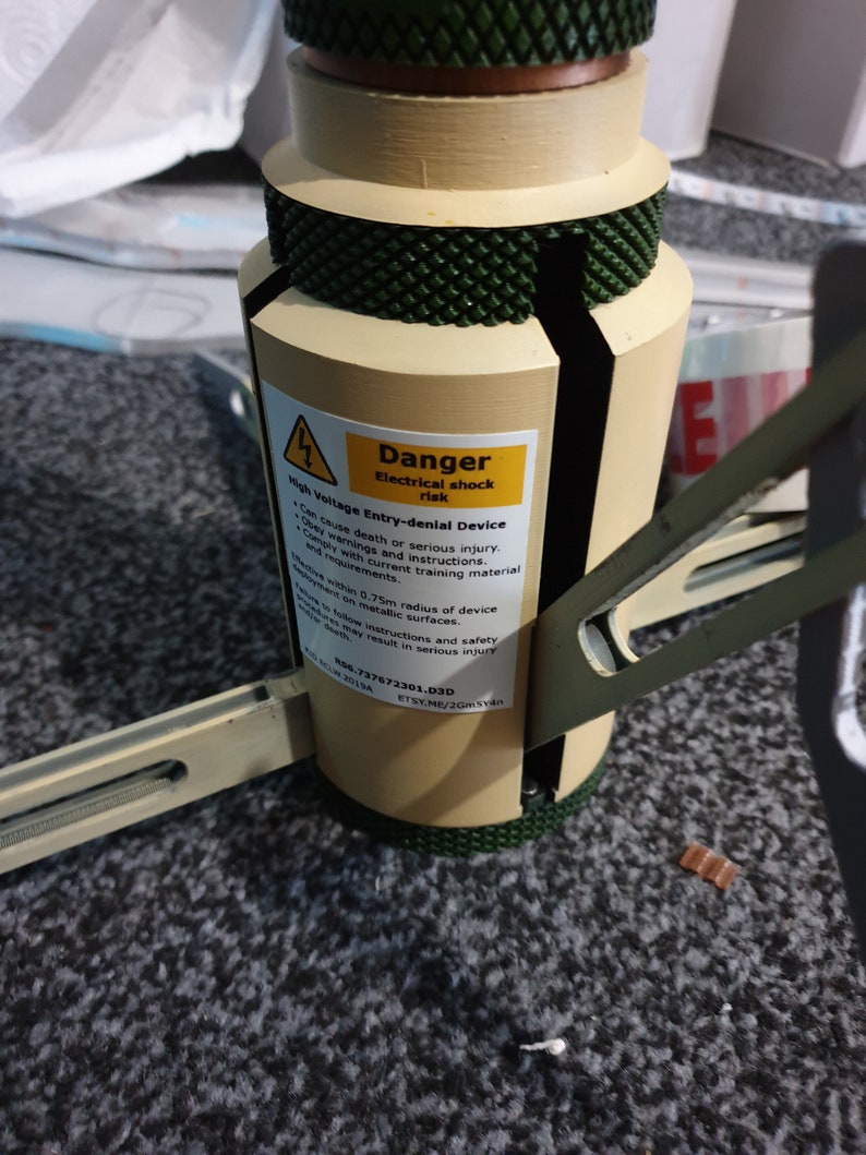 May include: A beige and green cylindrical device with a warning label that reads "Danger Electrical shock risk." The label also states "High Voltage Entry-denial Device." The device has a textured green top and base.