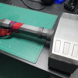 May include: A large, gray, metal hammer with a textured handle. The hammer has a rectangular head with three rectangular indentations on the top.