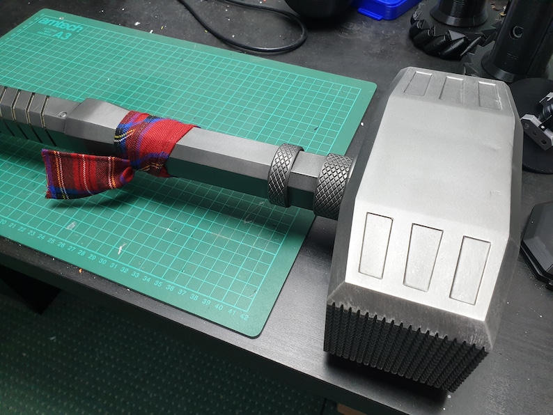 May include: A large, gray, metal hammer with a textured handle. The hammer has a rectangular head with three rectangular indentations on the top.