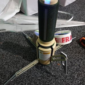 May include: A futuristic-looking device with a cylindrical, textured green body and a tan base. It has three angled legs and a glowing blue light at the top. A warning label is visible on the base. The device appears to be a prototype or a specialized tool.