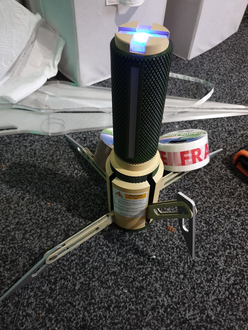 May include: A futuristic-looking device with a cylindrical, textured green body and a tan base. It has three angled legs and a glowing blue light at the top. A warning label is visible on the base. The device appears to be a prototype or a specialized tool.