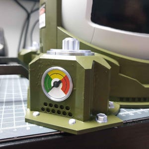 May include: A green and grey mechanical device with a round gauge featuring a black needle and a yellow, green, and red colour scale. The gauge is labelled with black numbers and a black arrow. The device has a silver knob and a series of small holes.