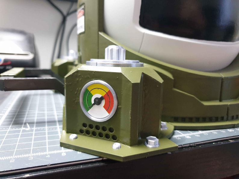 May include: A green and grey mechanical device with a round gauge featuring a black needle and a yellow, green, and red colour scale. The gauge is labelled with black numbers and a black arrow. The device has a silver knob and a series of small holes.