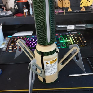 May include: A tan and green cylindrical device with four silver legs. The device has a diamond-patterned green top section and a white vertical stripe. A warning label is visible on the side.
