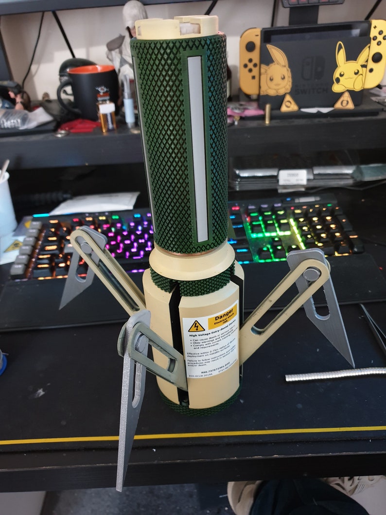 May include: A tan and green cylindrical device with four silver legs. The device has a diamond-patterned green top section and a white vertical stripe. A warning label is visible on the side.