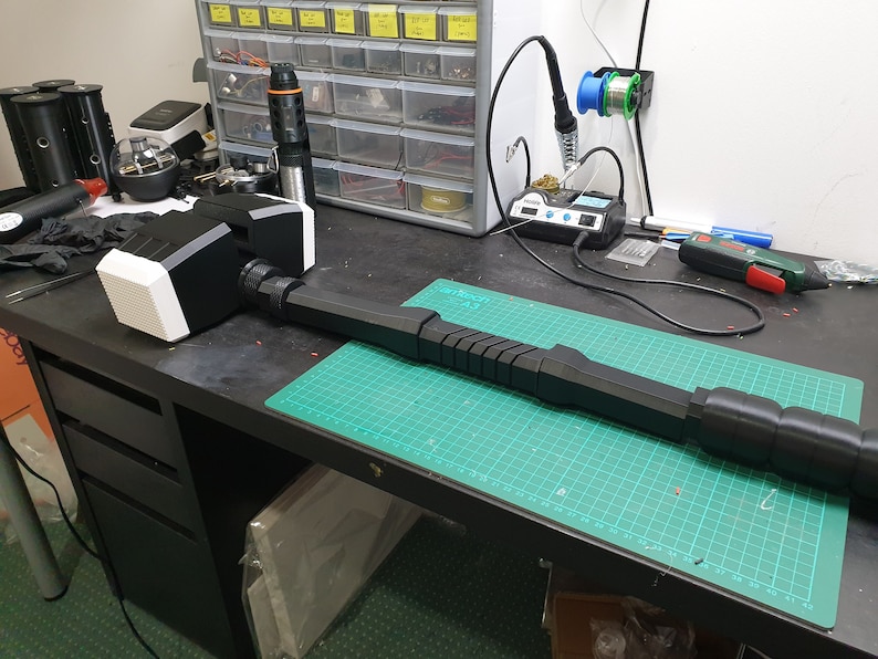 May include: A large, black and white prop hammer with a textured head and a long, faceted handle. The hammer is lying on a green cutting mat on a black workbench, suggesting a workshop setting. The hammer's head is white and black.