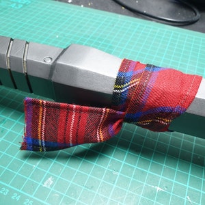 May include: A gray and black object wrapped with a red, blue, and yellow plaid fabric. The fabric is tied around the object and has a loose end.