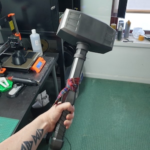 May include: A large, gray, 3D printed hammer with a black handle. The hammer is held by a person with a tattoo on their arm.