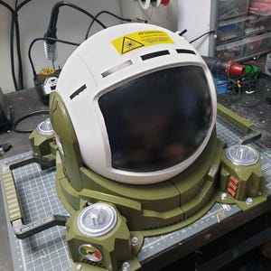 May include: A white astronaut helmet with a black visor and a green base. The helmet has a warning label that reads "Attention: Laser Hazard".