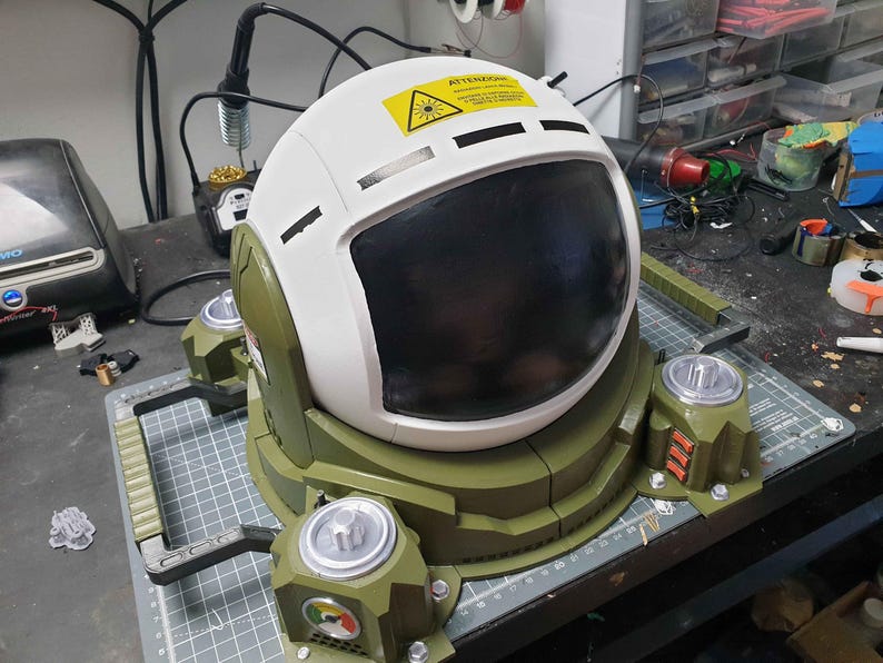 May include: A white astronaut helmet with a black visor and a green base. The helmet has a warning label that reads "Attention: Laser Hazard".