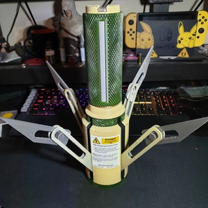 May include: A cylindrical device with a green textured body, a cream-colored base, and silver blades extending outwards. A warning label is visible on the side. The device is on a dark surface.