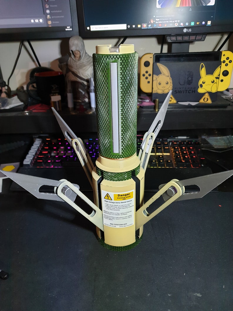 May include: A cylindrical device with a green textured body, a cream-colored base, and silver blades extending outwards. A warning label is visible on the side. The device is on a dark surface.